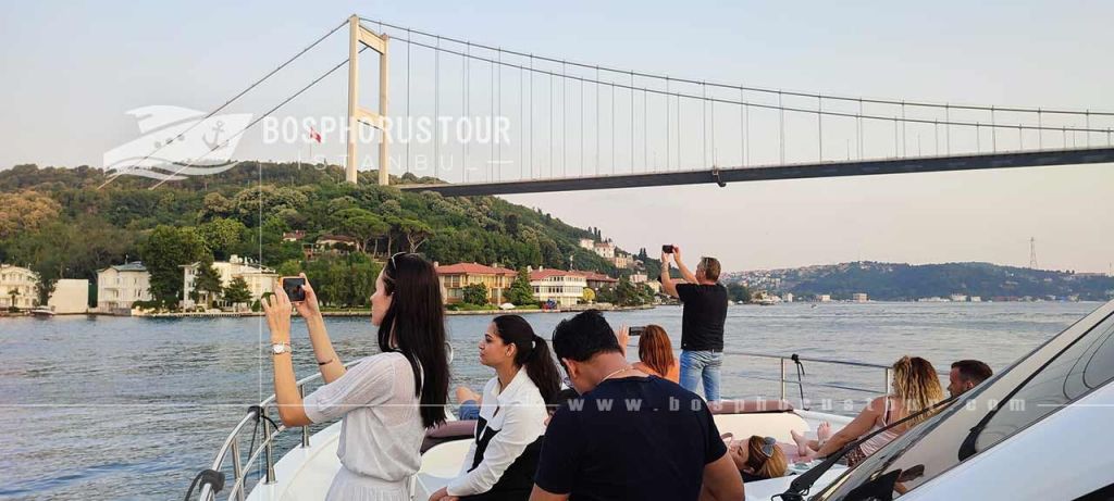 보트 투어에 참여한 사람들은 이스탄불의 보스포루스를 따라 가옥이 있는 다리와 경치 좋은 언덕의 사진을 찍습니다.