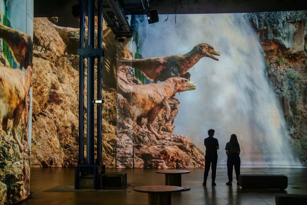 폭포가 있는 바위 배경에 공룡이 크게 투영된 방에 두 사람이 서 있습니다.