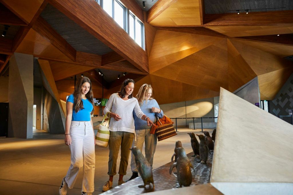세 명의 여성이 현대적인 목조 천장 건물 내부에 작은 펭귄 조각품을 전시하고 있습니다.