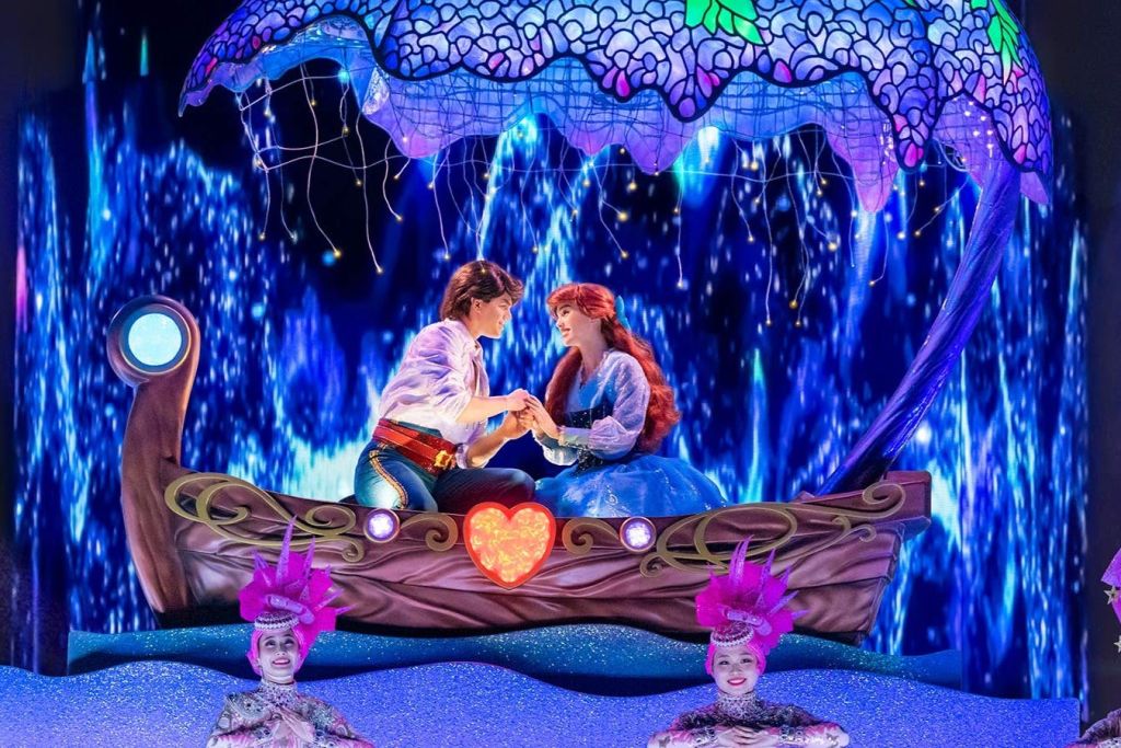 "인어공주" 의 캐릭터로 분장한 커플이 장식용 보트에서 손을 잡고 있으며, 아래에는 밝은 의상을 입은 공연자들이 있습니다.