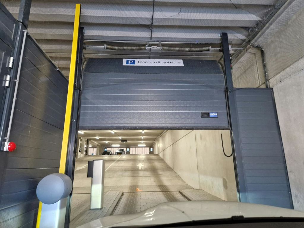 레오나르도 로얄 호텔의 부분적으로 개방된 셔터가 있는 지하 주차장 입구는 내부에 주차된 차량으로 이어지는 경사로를 보여줍니다.