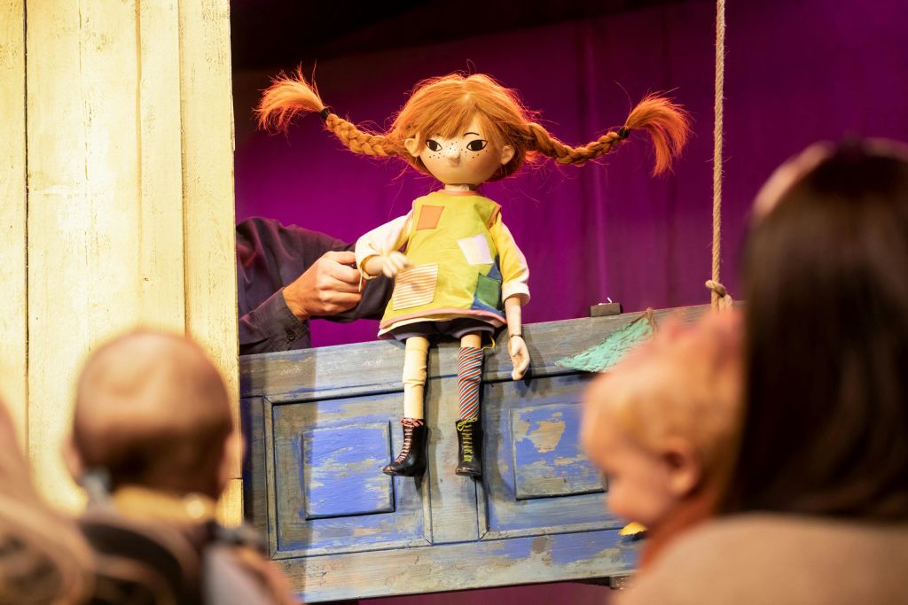 다채로운 색상의 옷차림으로 돼지꼬리가 달린 빨간 머리 인형은 푸른 나무 구조물 뒤에 있는 인형극단이 운영하며, 어린이들이 지켜보고 있습니다.