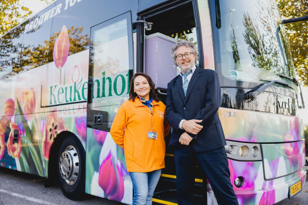 튤립 이미지로 장식된 화려한 버스 앞에 두 사람이 서 있습니다. 한 명은 오렌지색 재킷을 입고, 다른 한 명은 검은색 양복을 입습니다.