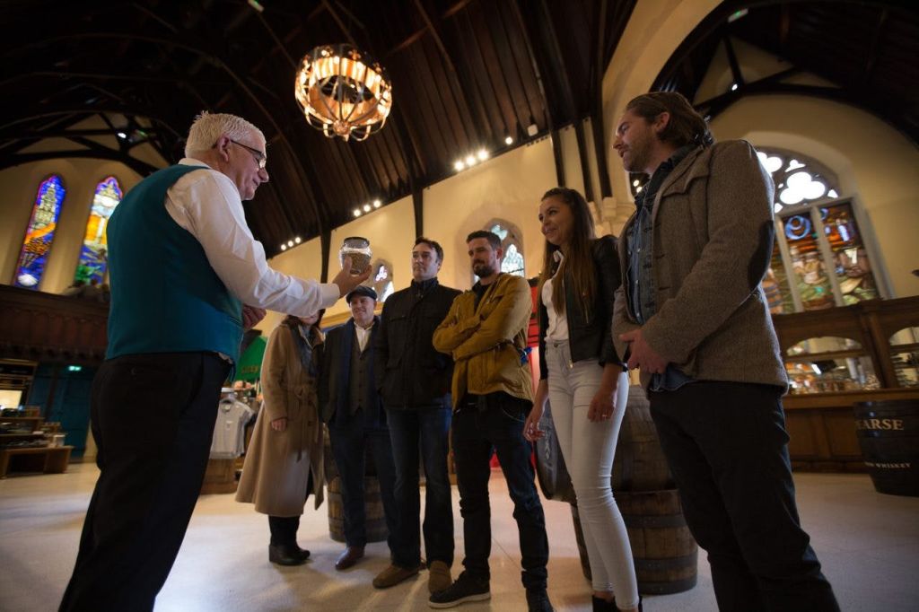 청록색 조끼를 입은 남성이 건물 내부에서 나무 기둥과 통을 든 다섯 명의 사람들에게 무언가를 설명하고 있습니다.