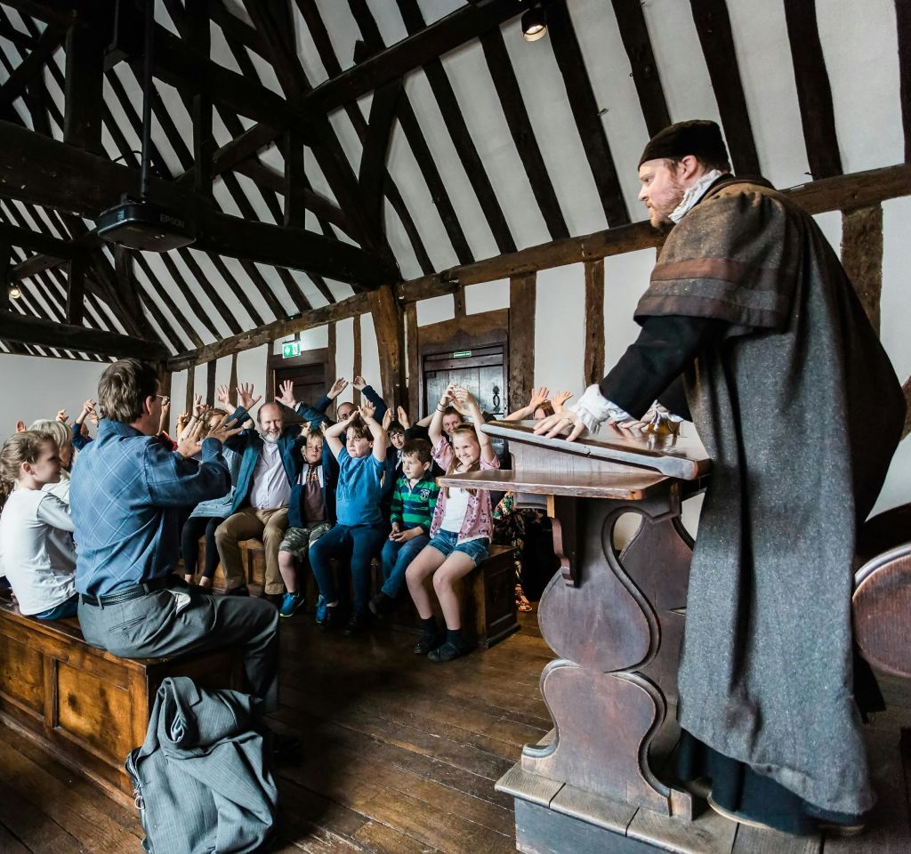 나무 기둥이 있는 방에서 벤치에 앉아 아이들을 강의하는 의상을 입은 남자. 아이들이 손을 들었습니다.