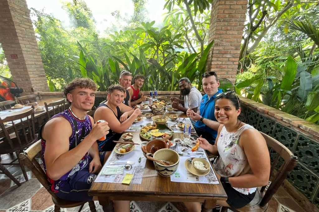 울창한 녹지와 벽돌 기둥으로 둘러싸인 야외에서 식사를 즐기는 6명의 사람들이 나무 테이블에 앉아 있습니다.