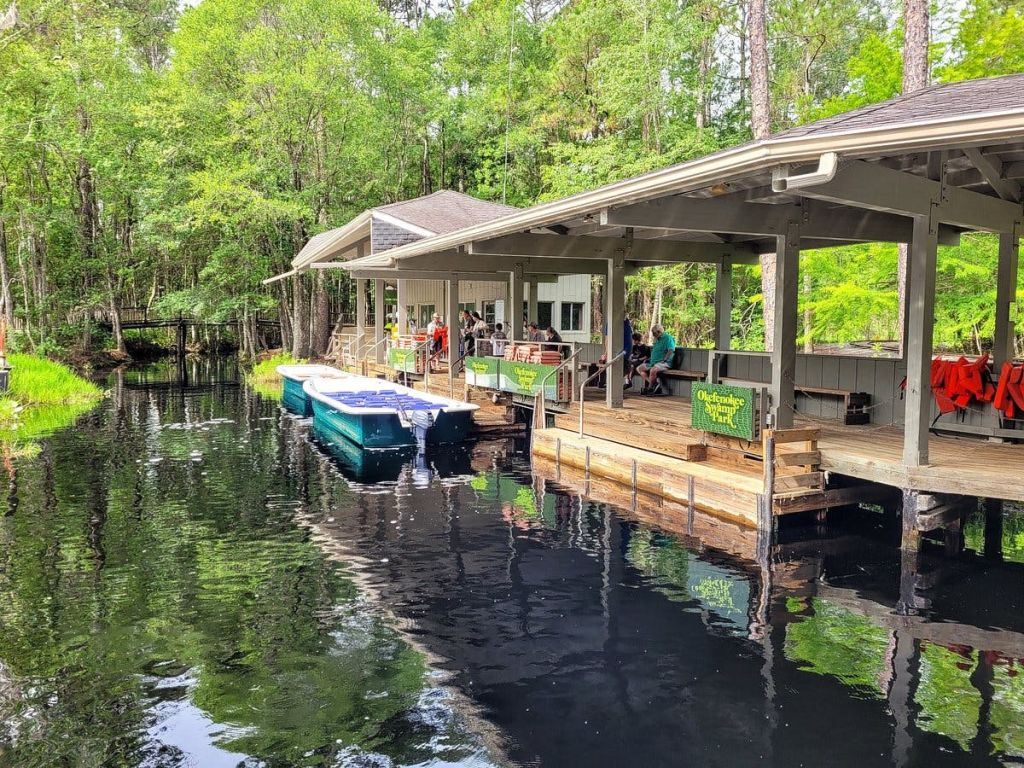 빽빽한 푸른 나무로 둘러싸인 잔잔한 강가에 목조 보트하우스가 자리하고 있습니다. 여러 척의 보트가 부두에 정박해 있습니다.