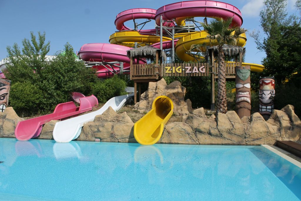 바위, 야자수, 녹지로 둘러싸인 투명한 수영장에서 끝나는 "지그재그" 라는 이름의 밝은 색상의 워터 슬라이드.