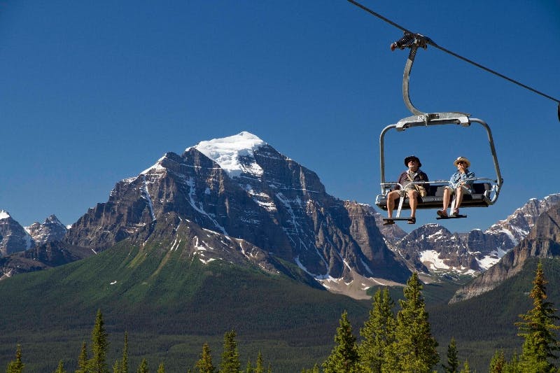 두 사람이 스키 리프트를 타고 눈 덮인 산과 맑고 푸른 하늘을 배경으로 숲을 가로지릅니다.