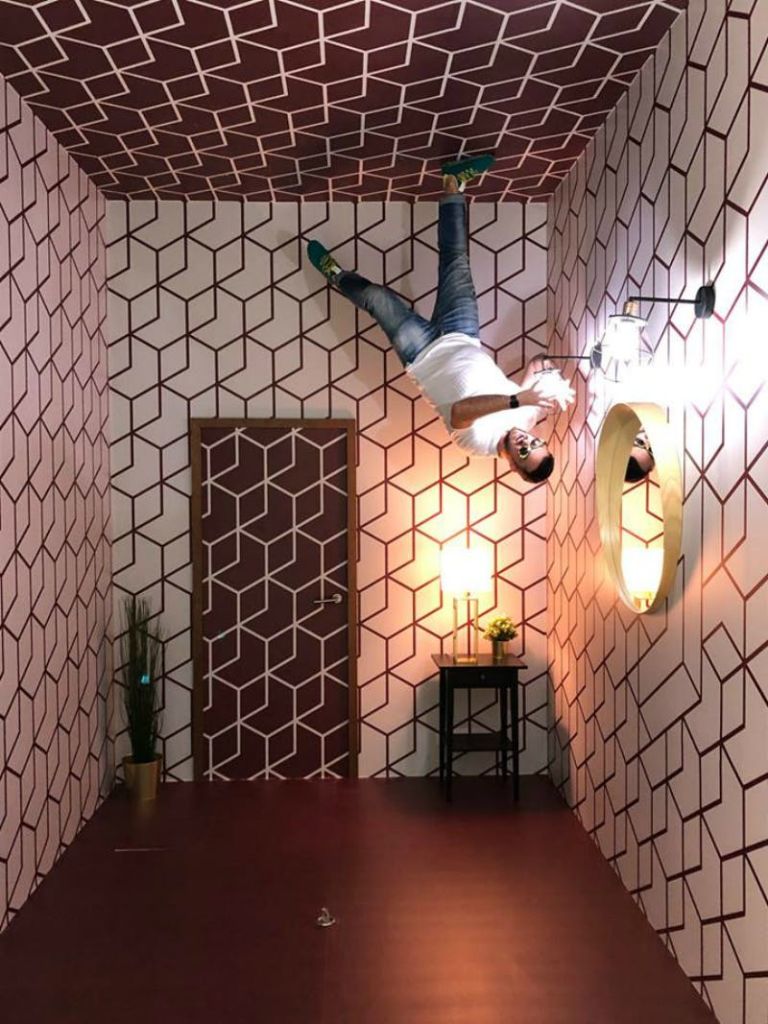 문, 거울, 램프가 있는 기하학적 패턴의 방 안에서 한 남성이 천장을 걷고 있는 듯한 모습으로 거꾸로 나타납니다.