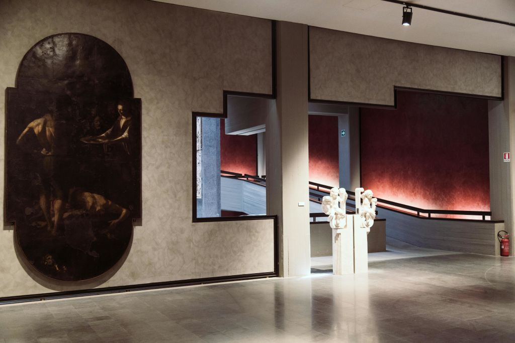 추상적인 벽 디자인, 왼쪽에 커다란 그림, 오른쪽 계단 근처에 조각된 동상이 있는 현대적인 박물관 인테리어.