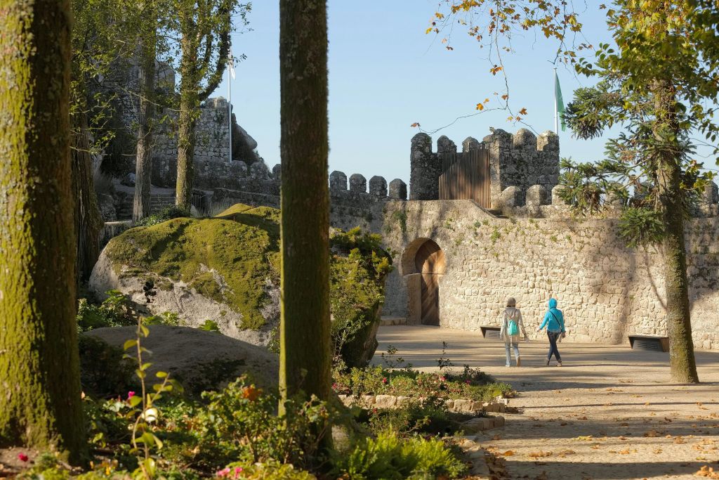화창한 날 녹음과 꽃으로 둘러싸인 고대 석조 성을 향해 걷는 두 사람.