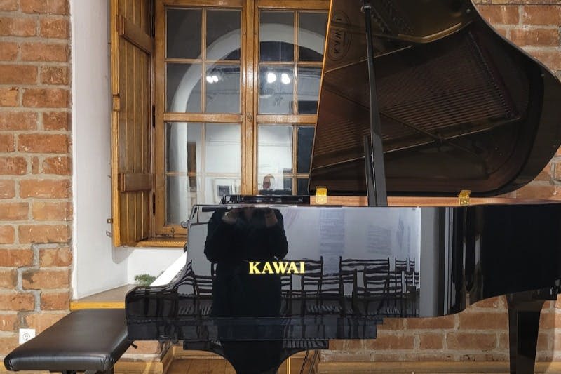 나무 창문과 벽돌 벽을 배경으로 사람과 의자가 비치는 검은색 카와이 그랜드 피아노.