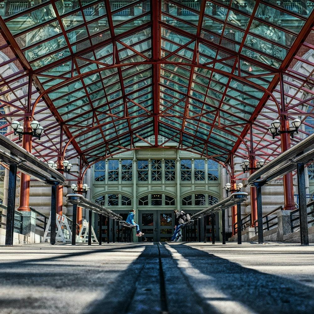 빨간색 금속 기둥이 있는 유리로 덮인 통로는 큰 입구로 이어지며, 가운데에는 빈티지 스타일의 램프와 두 사람이 있습니다.