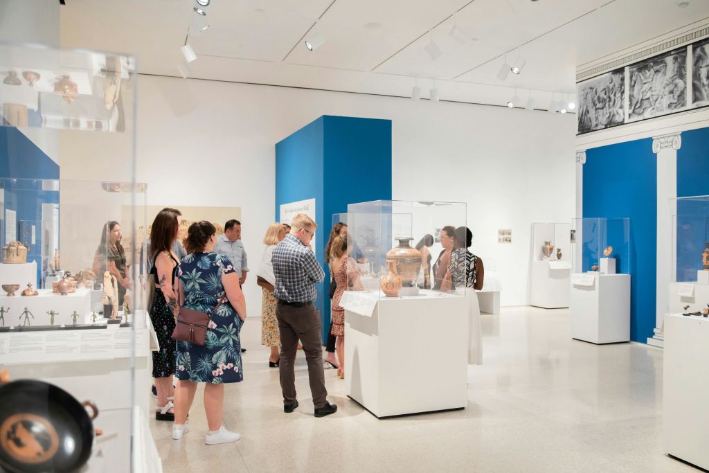 파란색 액센트 벽과 유리 케이스의 유물 전시가 있는 밝게 빛나는 박물관 갤러리의 전시물을 관찰하는 방문객.