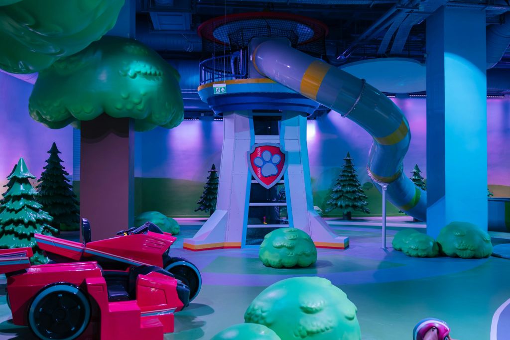 발 엠블럼, 나선형 슬라이드, 녹색 나무 장식, 빨간색 차량이 있는 탑이 있는 실내 놀이 공간.