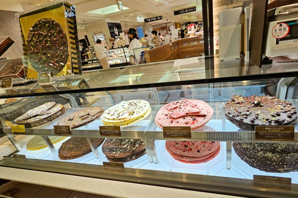 과자 가게에서 다양한 맛의 크고 화려한 초콜릿 디스크가 있는 디스플레이 케이스입니다. 고객과 카운터를 배경으로 합니다.