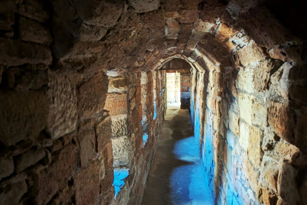 아치형 천장이 있는 좁은 석조 터널로, 작은 개구부와 끝의 출구에서 들어오는 자연 채광으로 조명을 받습니다.