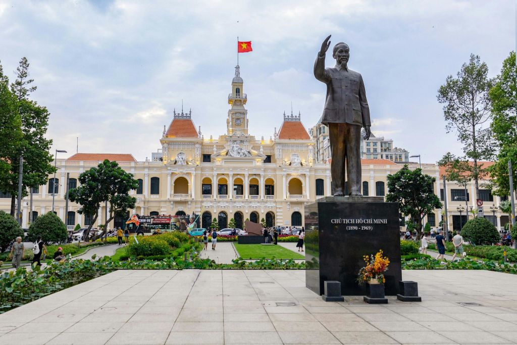 나무와 사람으로 둘러싸인 베트남 국기가 그려진 커다란 화려한 건물 앞에 있는 호치민 동상.