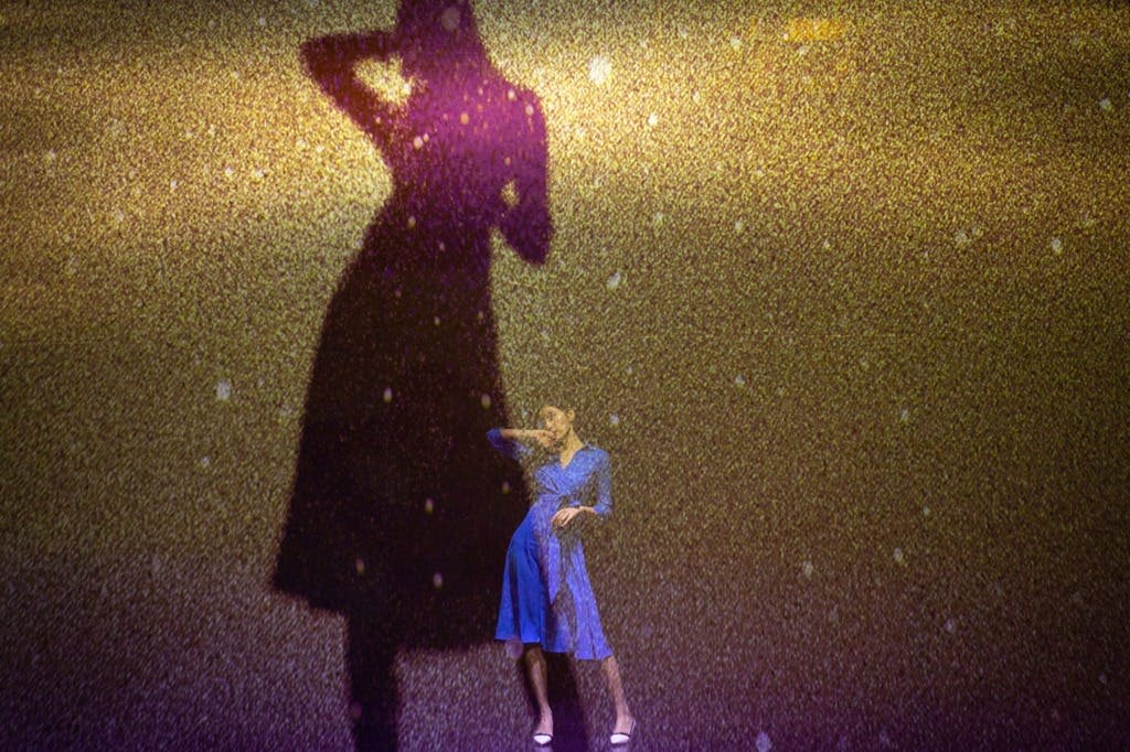 파란색 의상을 입은 사람이 그림자가 두드러지게 보이는 질감의 황금색 배경 앞에서 포즈를 취하고 있습니다.