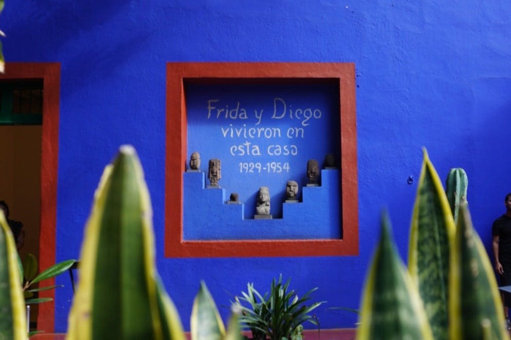"Frida y Diego vivieron en esta casa 1929-1954" 라는 스페인어로 작은 조각상과 텍스트가 표시된 빨간색 프레임의 틈새 시장이있는 파란색 벽.