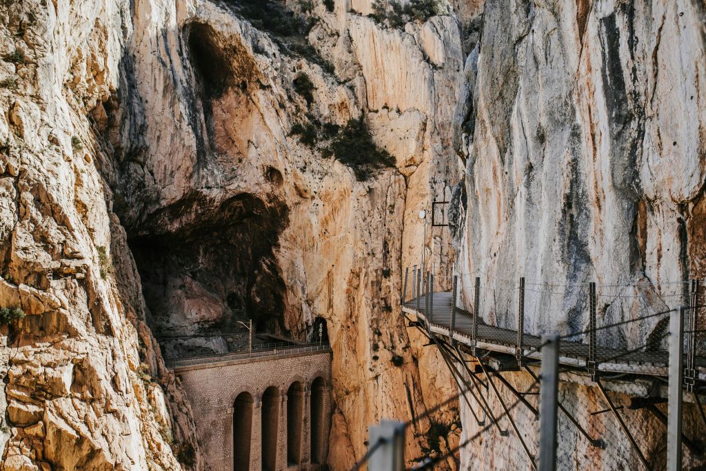 난간이 있는 좁은 산책로가 가파른 바위 협곡을 따라 이어지며, 아래에는 아치형 다리가 보입니다.