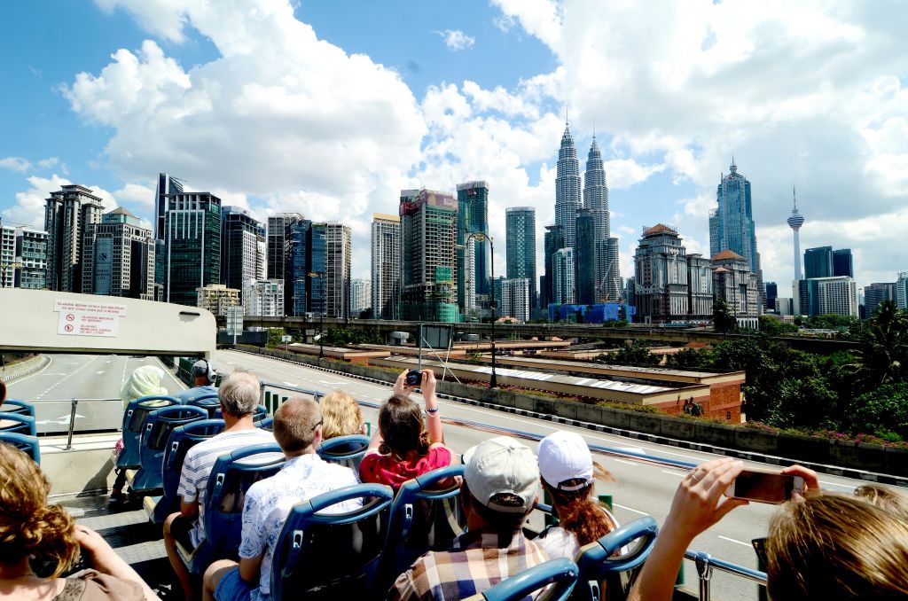 현대적인 고층 건물과 밝고 푸른 하늘 아래 페트로나스 타워가 있는 도시 풍경이 내려다보이는 2층 버스 투어에 참여한 사람들.