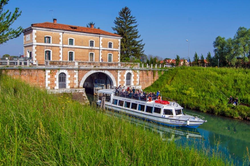 맑은 하늘 아래 푸른 잔디와 나무로 둘러싸인 아치형 개구부가 있는 벽돌 건물 옆으로 승객이 탄 보트가 터널로 들어오고 있습니다.