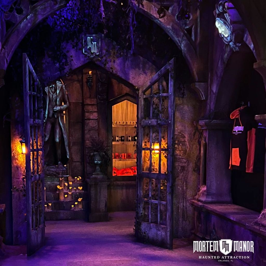 탁 트인 철제 문, 고딕 양식의 장식, 기이한 동상, 조명이 켜진 촛불이 있는 유령의 입구에 희미한 조명을 비춥니다. 표지판에는 "Mortem Manor Haunted Attraction" 이라고 쓰여 있습니다.
