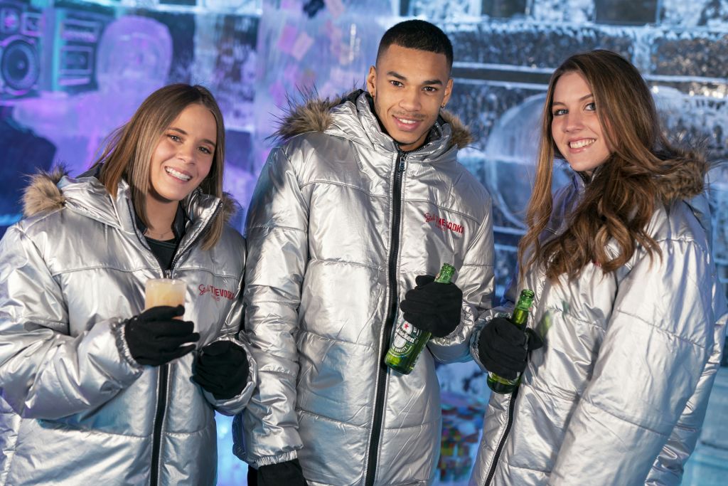 실버 재킷을 입은 세 사람이 얼음으로 뒤덮인 배경을 배경으로 가까이 서서 음료를 들고 미소를 짓고 있습니다.