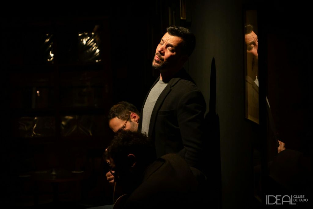 어두운 블레이저를 입은 한 남성이 반사 표면이 흐릿하게 비치는 벽에 맞서고 있으며, 그 뒤 부분적으로 보이는 또 다른 남성이 있습니다.