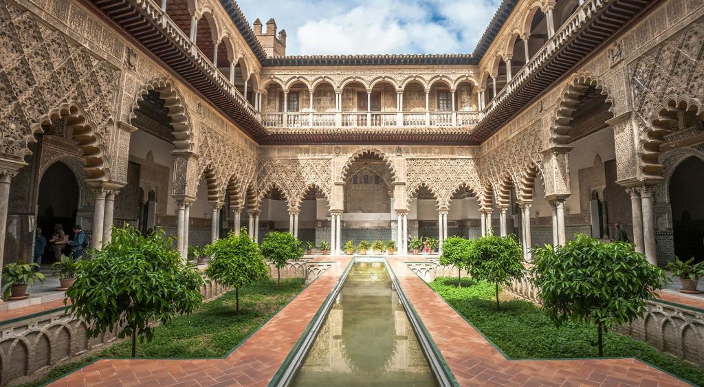 화려한 아치, 반사되는 긴 중앙 수영장, 부분적으로 흐린 하늘 아래 녹음이 우거진 마당.