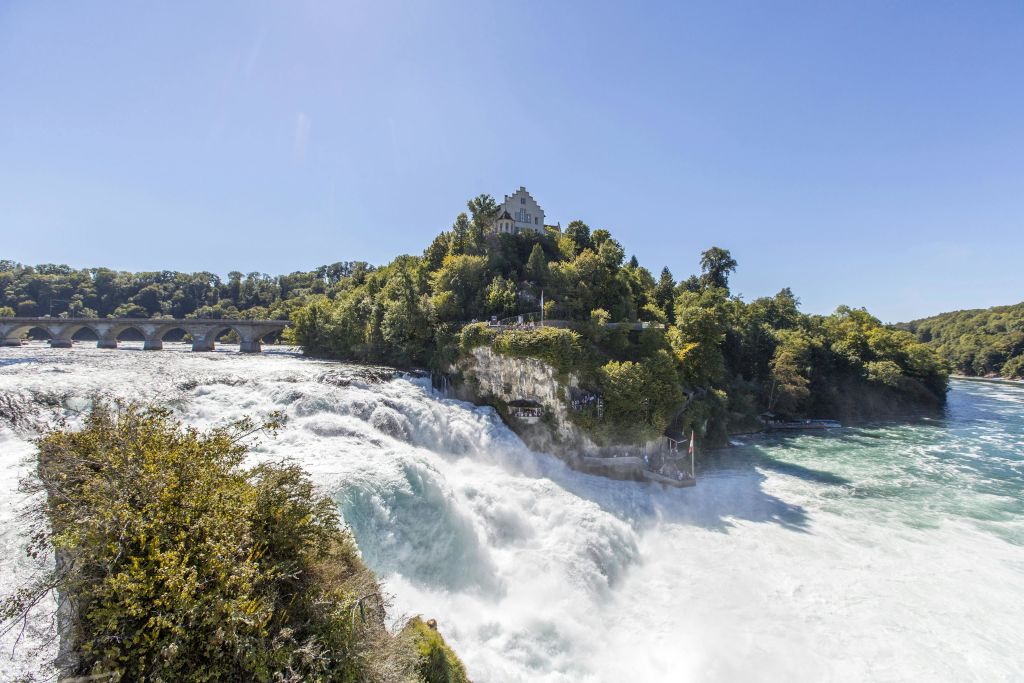 나무로 덮인 언덕과 맑고 푸른 하늘 아래 꼭대기에 성 모양의 건물이 있는 바위를 따라 계단식으로 이어지는 대형 폭포.