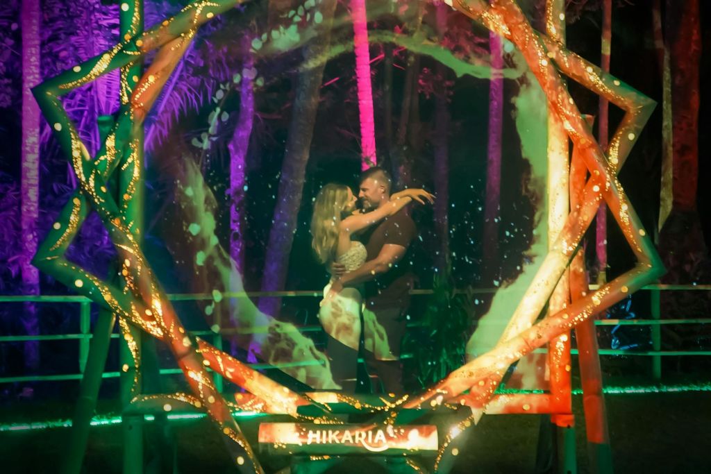 밤에는 화려하고 빛나는 나무로 둘러싸인 "HIKARIA" 표지판이 있는 불빛이 들어오는 자연을 테마로 한 아치 아래 부부가 포옹하고 있습니다.