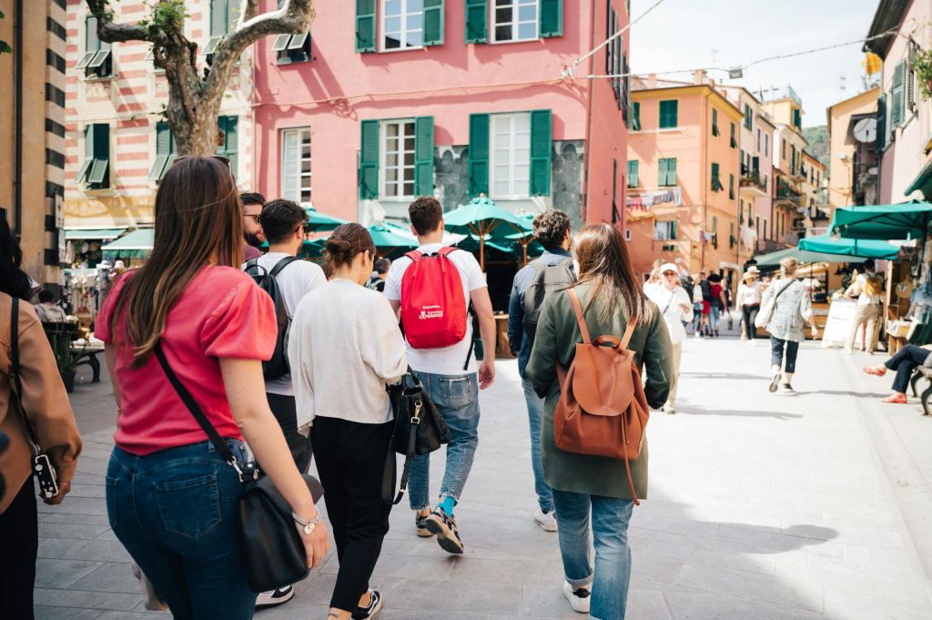 한 무리의 사람들이 분홍색과 주황색 건물과 녹색 창문 셔터가 있는 햇살이 내리쬐는 다채로운 광장을 거닐고 있습니다.