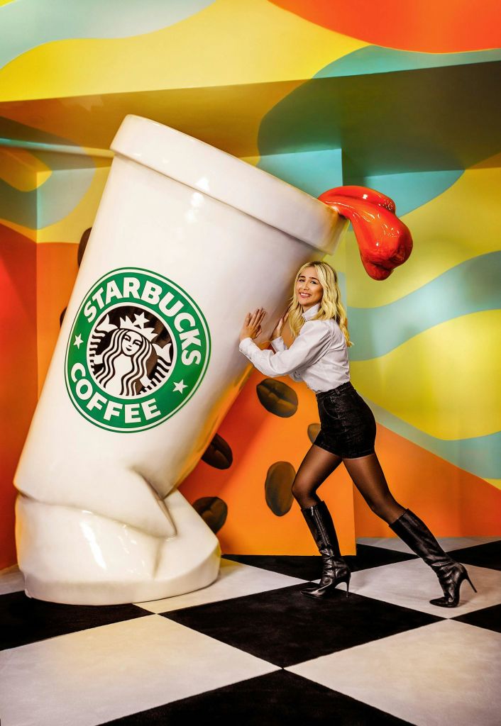 화려하고 추상적인 배경 속에서 오버사이즈 스타벅스 커피 컵을 들고 포즈를 취하는 여성. 빨간색 액체를 엎지르는 컵.