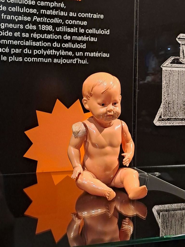 어깨가 손상된 마모된 알몸 플라스틱 아기 인형이 프랑스어 텍스트와 삽화와 함께 검은색 배경에 표시됩니다.