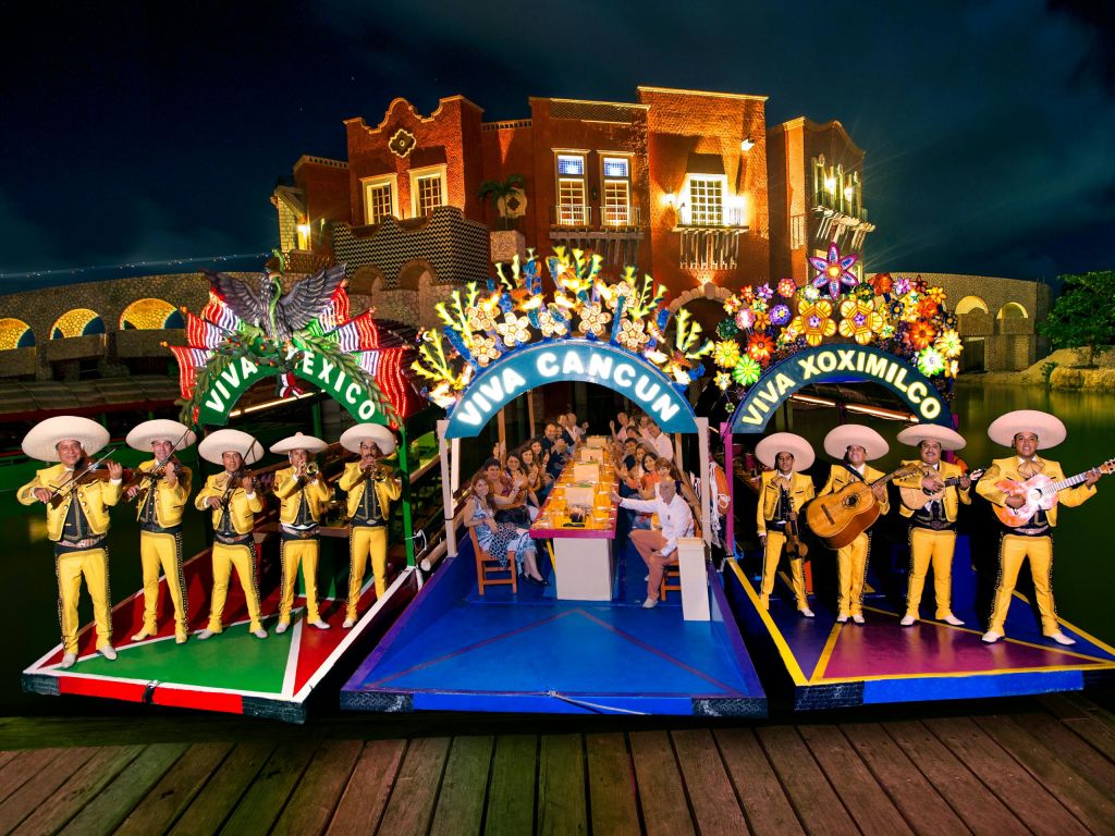 화려한 색상의 두 마리아치 밴드가 밤에 장식된 보트에서 공연을 펼치고, 사람들은 식사를 하며 환호합니다. 조명이 켜진 건물이 배경입니다.
