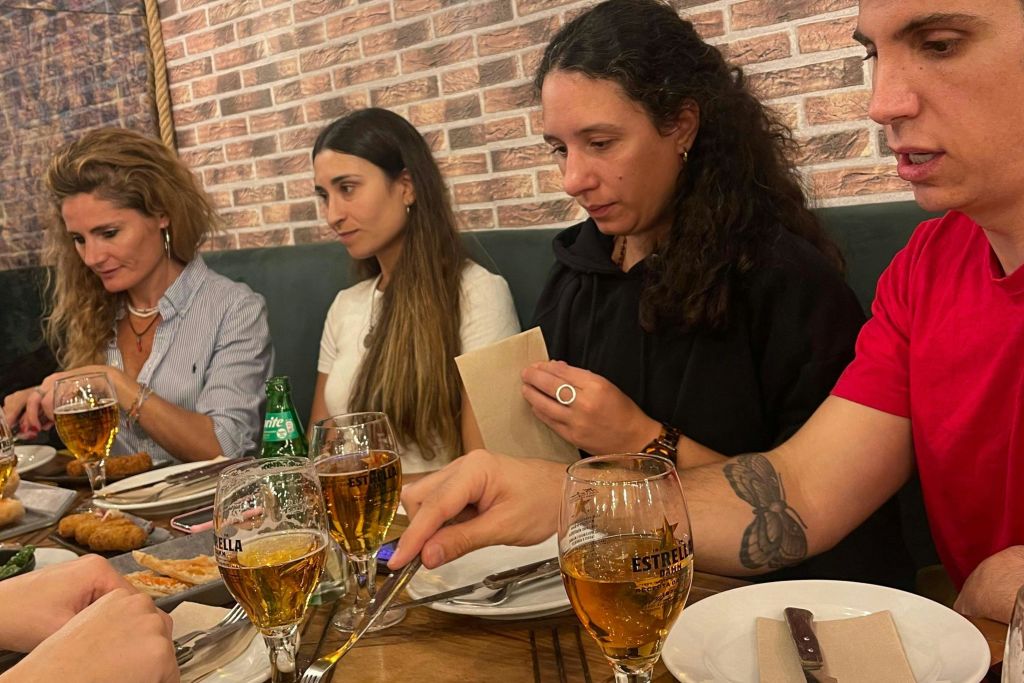 벽돌 벽을 배경으로 에스트렐라 맥주잔이 놓인 테이블에 세 사람이 앉아 있습니다. 한 사람이 식기류를 찾으려고 손을 뻗습니다.