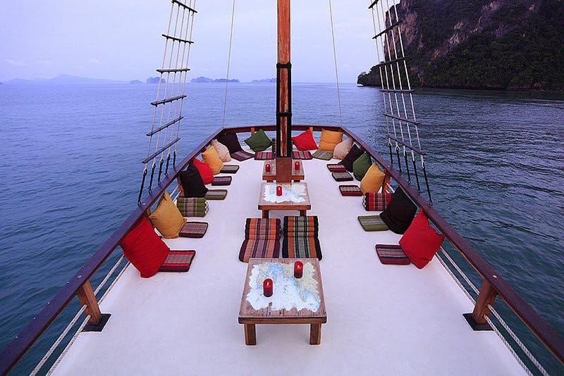 쿠션이 있는 좌석, 낮은 테이블, 촛불이 놓인 보트 데크, 멀리 떨어진 섬과 절벽을 배경으로 잔잔한 바다에서 요트를 즐겨보세요.