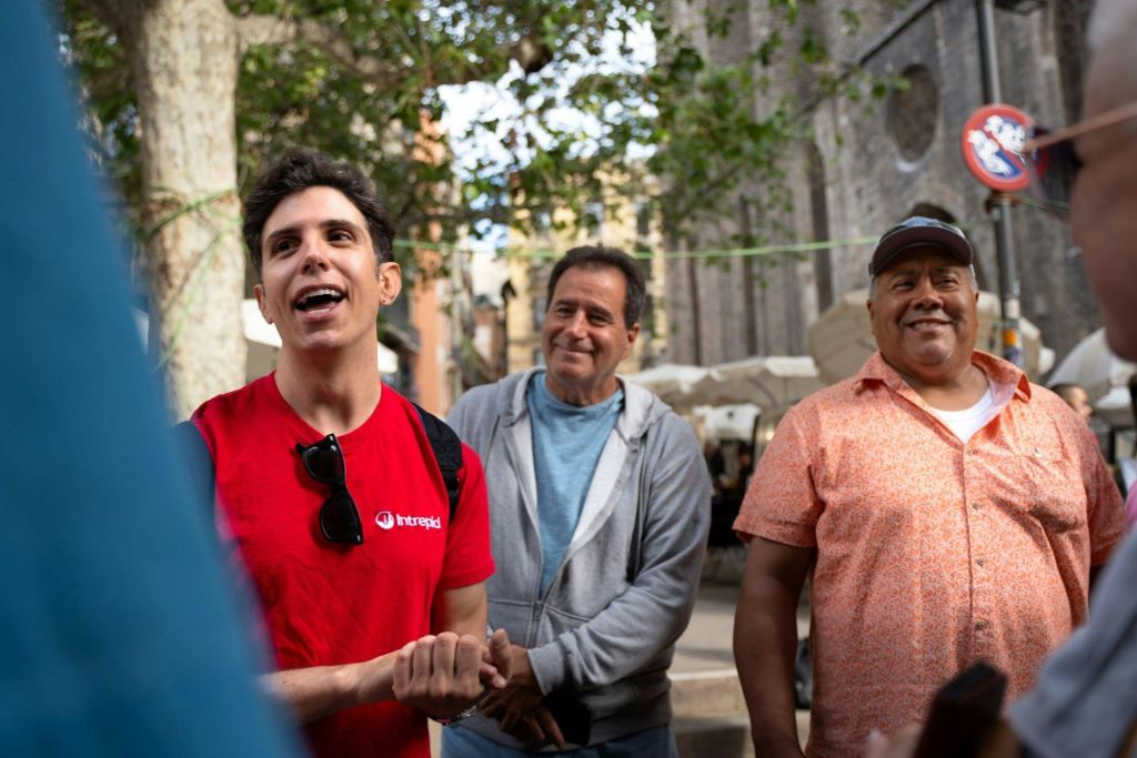 빨간 셔츠를 입은 남성이 야외에서 소규모 그룹과 대화하는 동안 제스처를 취하고, 다른 남성이 그의 뒤에 서서 미소를 짓고 있습니다. 나무와 건물이 배경입니다.