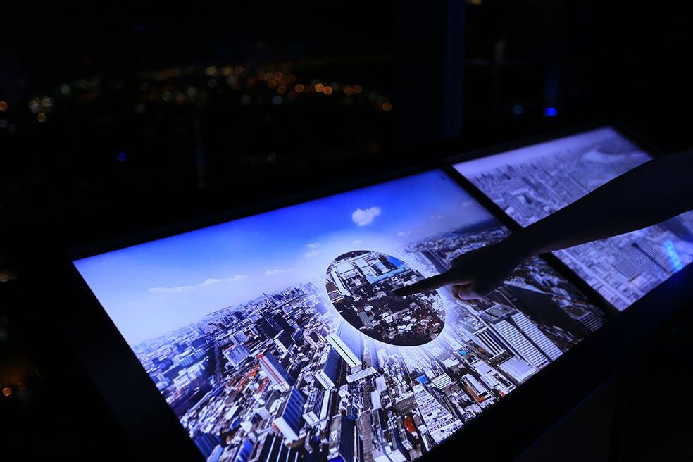 한 사람이 야간에 도시의 공중 전망을 보여주는 터치스크린과 상호 작용하며, 원형으로 확대된 섹션이 있습니다.