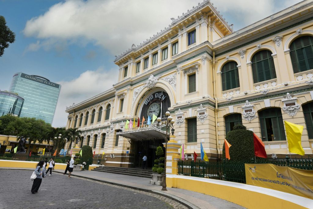 화려한 노란색 건물에 아치형 출입구, 시계, 여러 개의 깃발이 있습니다. 앞에 두 사람이 있습니다. 위쪽은 흐린 하늘입니다.