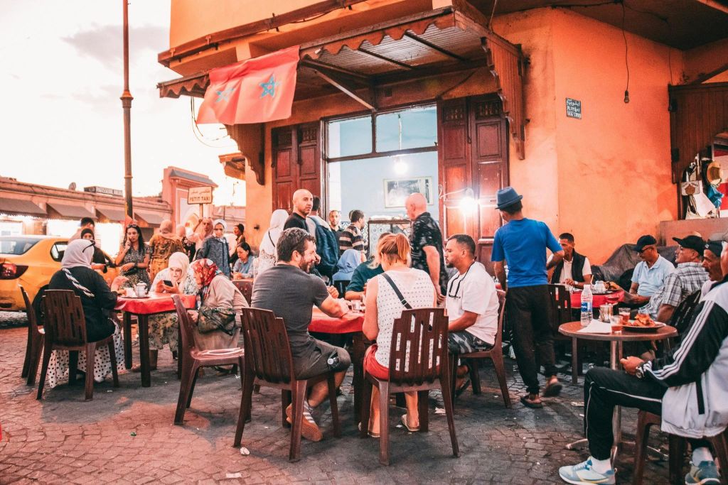 나무 가구, 주황색 벽, 모로코 국기가 있는 야외 카페에 앉아 대화를 나누는 사람들.