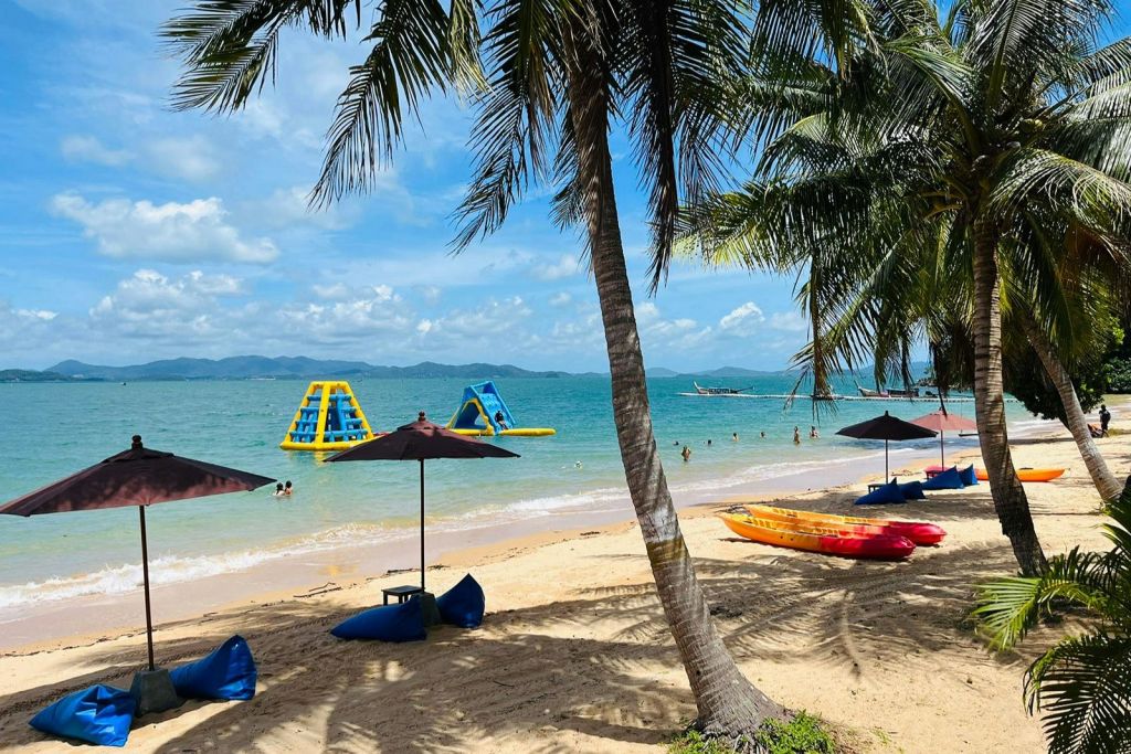 야자수, 카약, 파라솔이 있는 모래 해변. 맑고 푸른 바다에서 팽창식 구조물 옆을 헤엄치는 사람들.