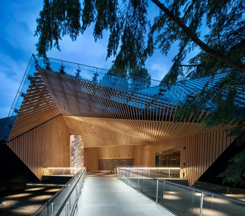 나무 판자와 유리 난간이 있는 현대적인 건물로, 나무 사이에 설치되어 있으며 해질 무렵에 조명을 받습니다.