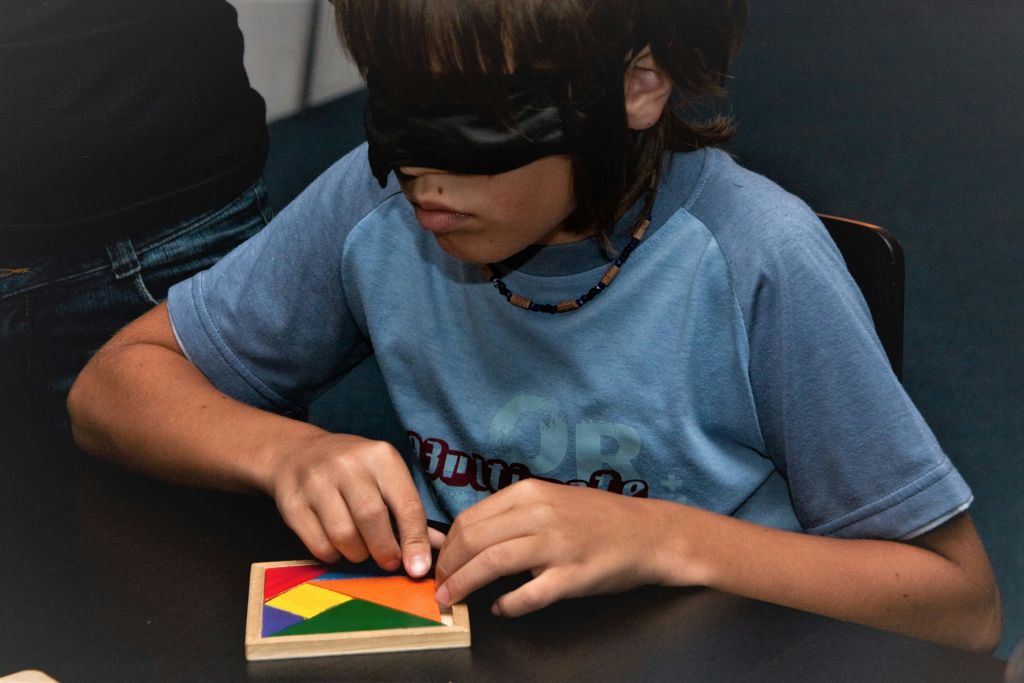 파란색 셔츠와 구슬 목걸이를 입은 눈가리개를 한 어린이가 터치를 사용하여 작은 나무 보드에 다채로운 모양을 배열합니다.