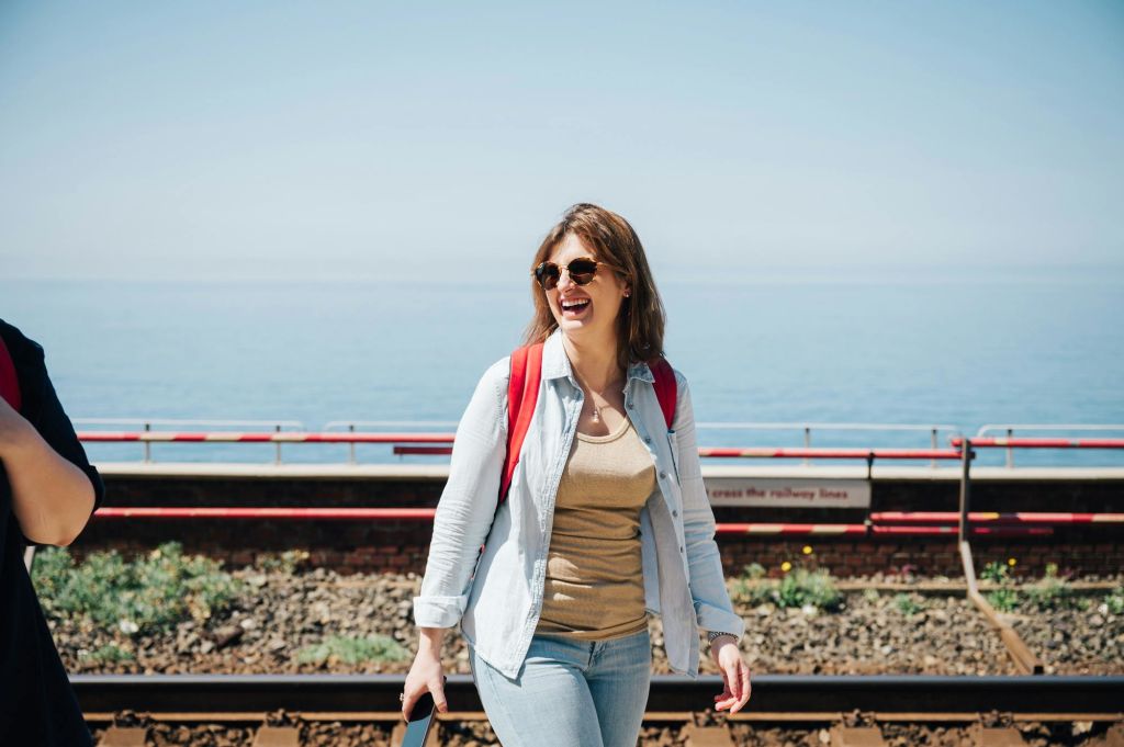 선글라스를 끼고 빨간색 배낭을 메고 바다가 보이는 기차 트랙 근처를 걷는 여성.