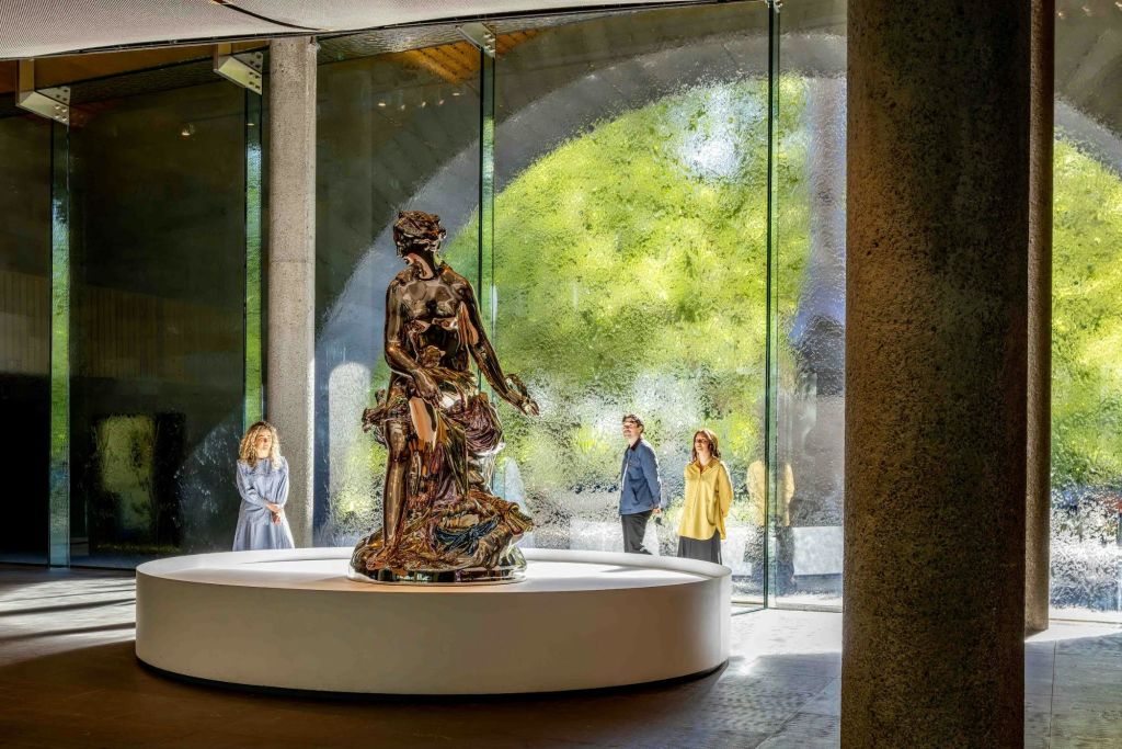 유리 벽이 있는 실내 공간의 원형 받침대에 사람들이 관찰하고 녹지를 배경으로 전시된 동상입니다.