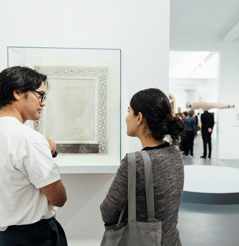 두 사람이 갤러리에 서서 액자에 담긴 작품에 대해 토론하고 있으며, 여러 사람과 다른 작품이 배경으로 보입니다.